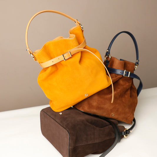 Three bucket Danilos Bags in suede leather in yellow, brown, and dark brown on a white surface with a beige background.