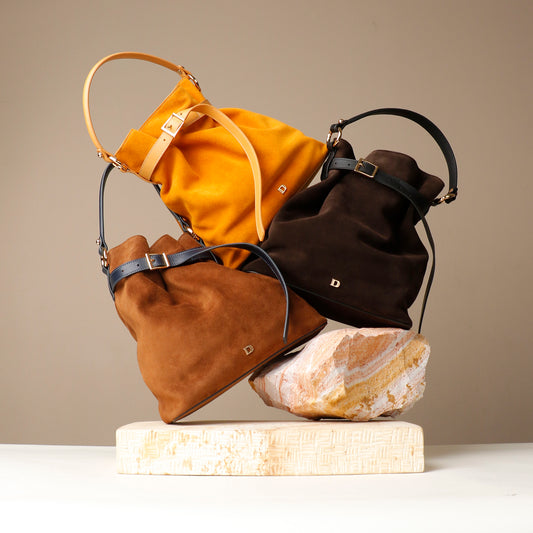 Three bucket Danilos bags in yellow, brown, and dark brown on a stone block with a beige background