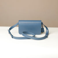 Blue leather crossbody bag with a strap on a white surface and beige background