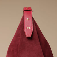 close up of a red suede leather danilos  bag with a strap featuring a gold 'D' on a beige background