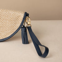 Woven raffia Agnes clutch with navy leather strap and tassel on a beige background