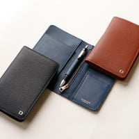 Three danilos travel pro passport cases in black, brown, and blue leather colors, displayed open with a pen resting on the black case, set against a neutral background.