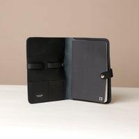 Front view of an open black Danilos pebble leather inskspirator with a clip closure, a notebook inside, and organized pouches for pens on a beige background