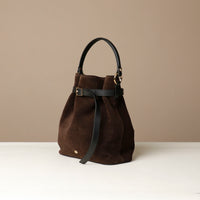 side of a Brown suede bucket Danilos bag with black handle and strap on a beige background