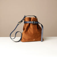 Brown suede leather bucket Danilos bag with a long black strap to crossbody it on a beige background