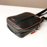 close up view of a open zipper expandable danilos black and brown crossbody dylan bag over a beige background